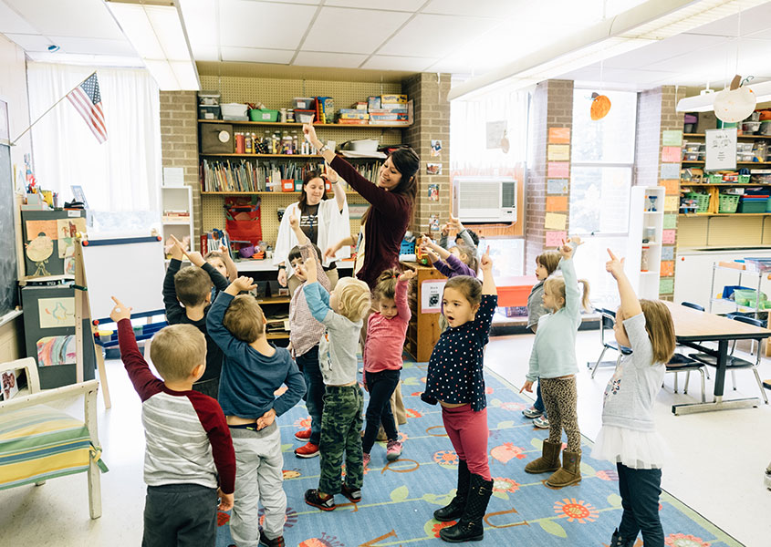 Preschool children and teacher in a classroom all pointing upwards