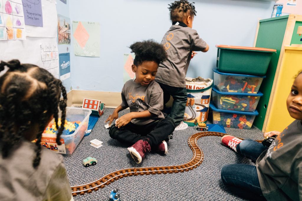 Image: Several young children sit together on the floor, each playing with a toy of their choice.