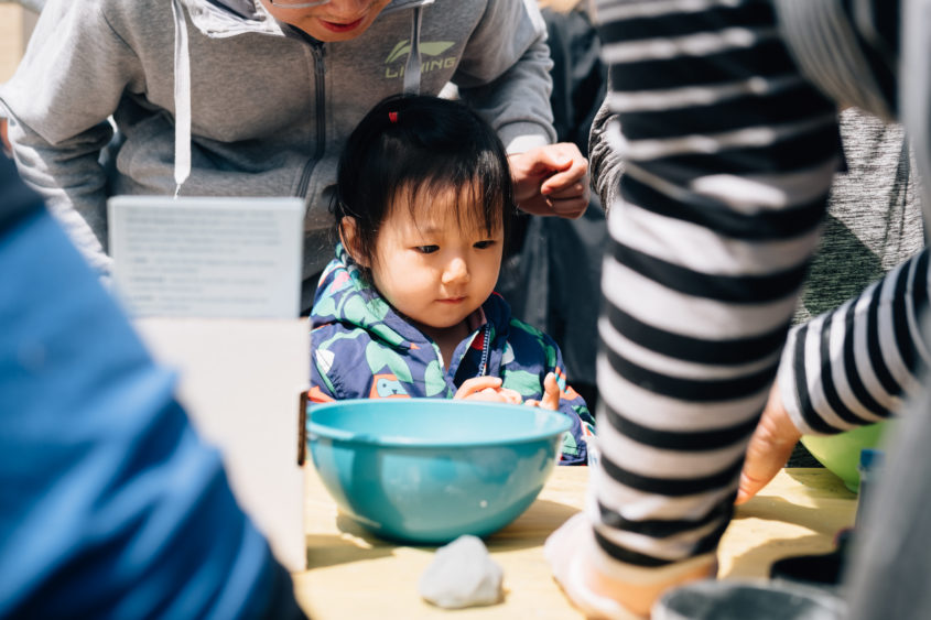 Small child with caregiver stands at table with gray clay.