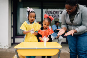 Two girls and caregiver playing with white slime in front of barbershop.