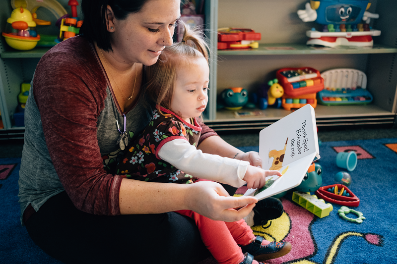 Caregiver and infant sit and read a book together. Interested in the beautiful yellow color and words, the baby points a finger at the page.