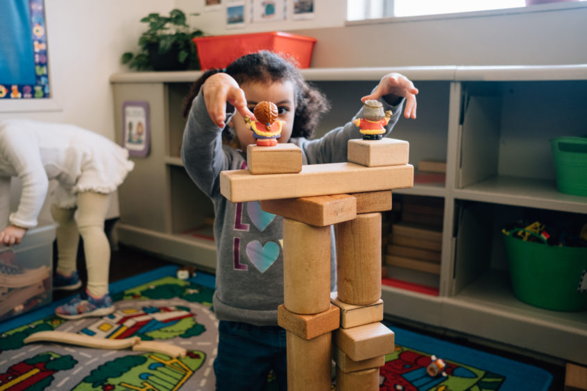 Image: A young child carefully stacks blocks into a tower as another young child digs around through toys in the background.