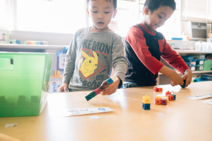 Image: Two young children stand together at a table, both playing with small colorful blocks.