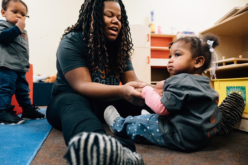 Image: An early learning professional sits on the ground, interacting with two young children.