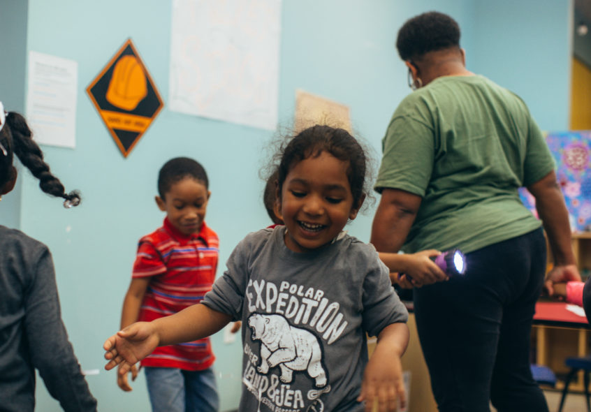 Three young children run around a room while giggling and having fun. One shines a flashlight as a caregiver stands beside them.