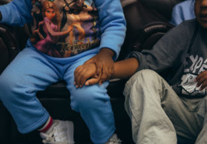 Two young children sit together; one grasps the wrist of the other.