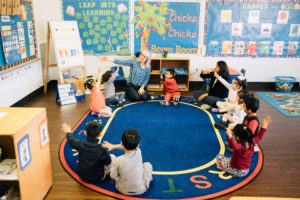 Young children sit in a circle on a rug as the lead teacher points her hand toward a visual aid.