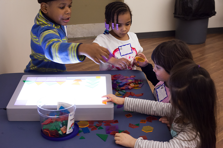 Picture: Four young children stand around a table, playing with translucent shapes on a light board.