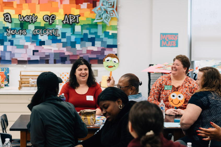 Image: Early childhood professionals join together as a group, with one woman holding up a happy green face on a paper plate.