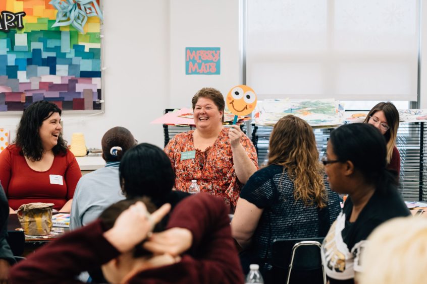 Image: Early childhood professionals sit together, all looking at a professional who is holding up a smiley face on a stick.
