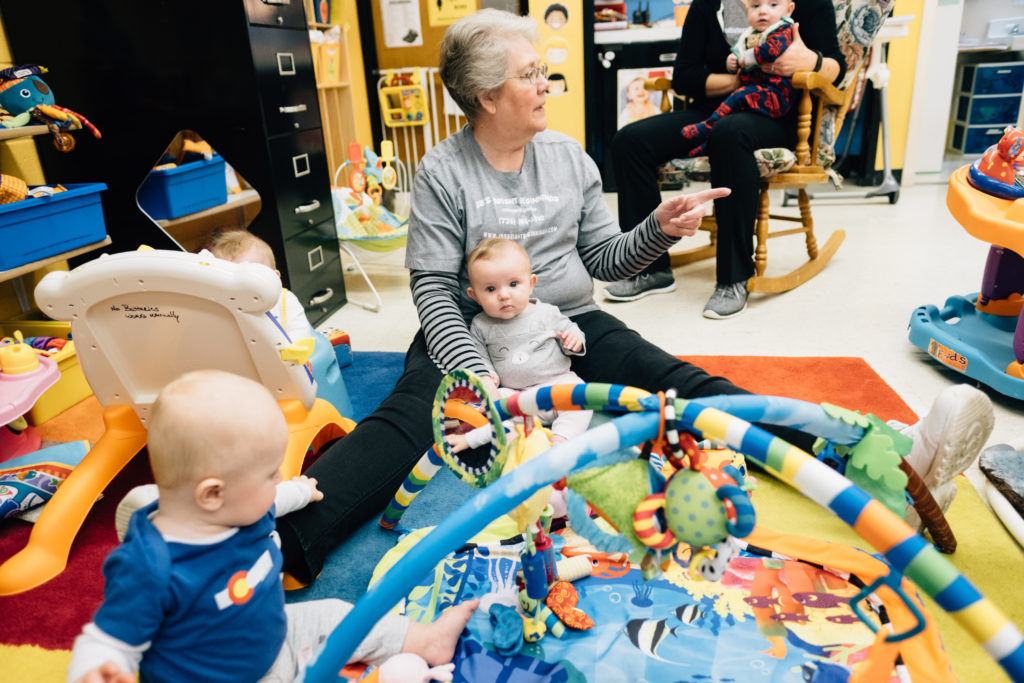 Image: An older child care provider sits together with two babies as another provider sits in a chair holding a baby.