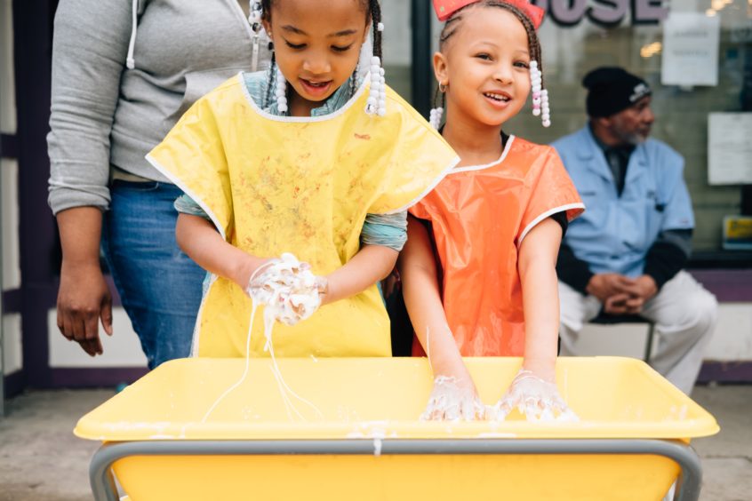 Image: Two young children stand together with their hands reaching in a box of mushy goo,