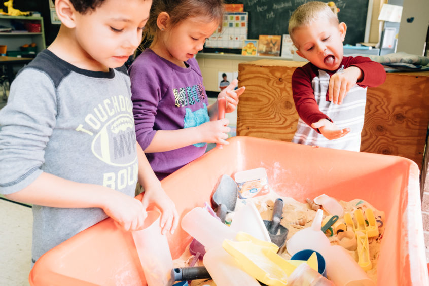 Image: Three young children play together in a raised bin with sand and toys in it.