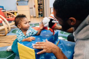 A baby and adult male caregiver share a close interaction as the caregiver interacts puppets an orange and black toy.