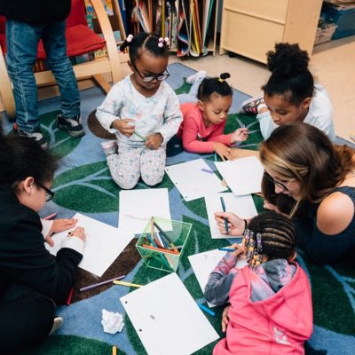 A group of young children sit together in a circle on the ground, writing on pieces of paper and talking to an early learning professional.