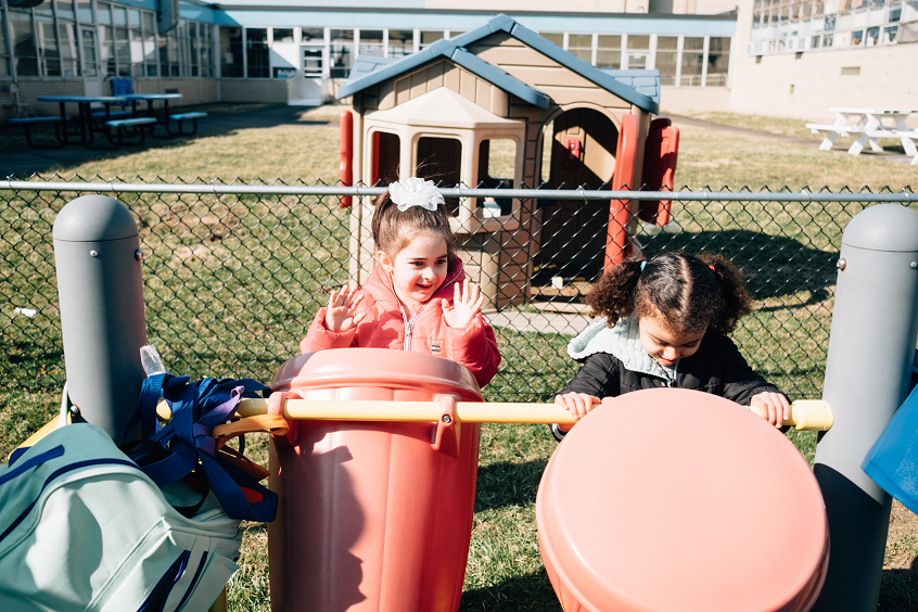 Image: Two young children play together at a playground, each rocking and tapping on big, moving bins.