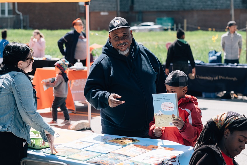 Image: A male caregiver stands beside his young child, as the child picks out books from the Imagination Library stand at the Homewood Block Party.