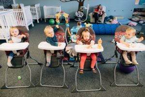Image: Four babies sit in a line of highchairs, looking all around the room happily.