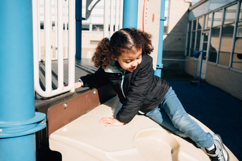 Image: A young child slides down a play obstacle, with their hands holding them up gently.