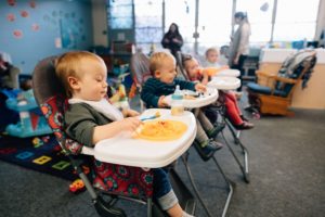 Image: Young children sit in highchairs, eating food on plates in front of them.