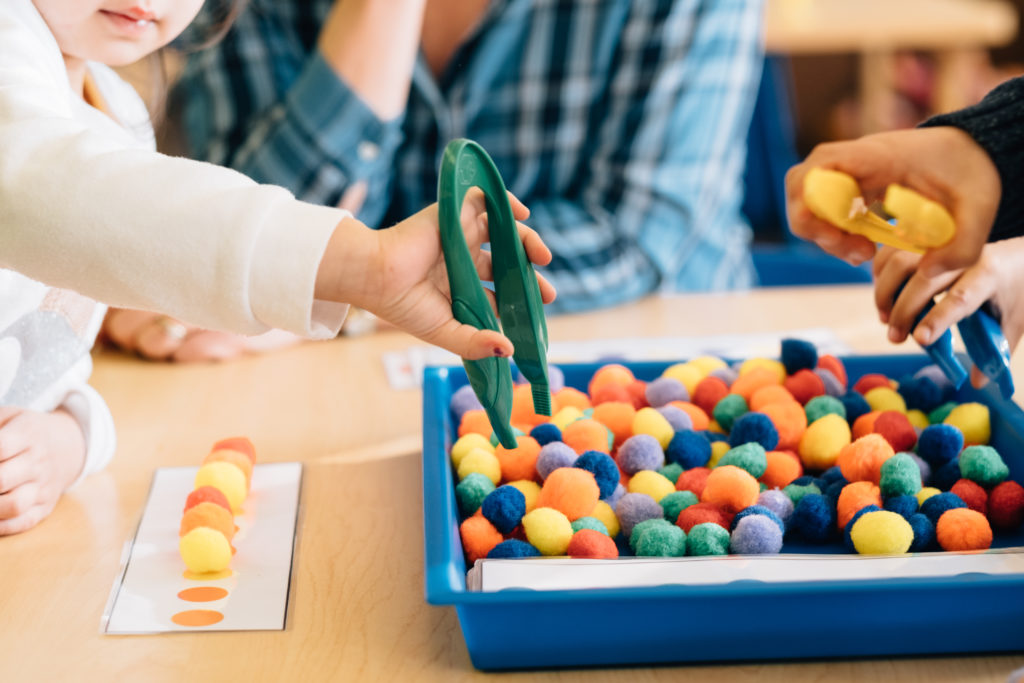Image: A young child sits beside an early learning professional, holding tongs to grab colorful cotton balls for a matching activity.