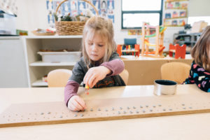 Image: A young child plays with a small object at a table, hovering it above a wooden board.