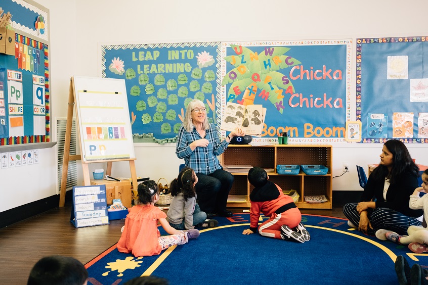Image: An early learning professional sits facing a group of young children, reading a picture book aloud while smiling.