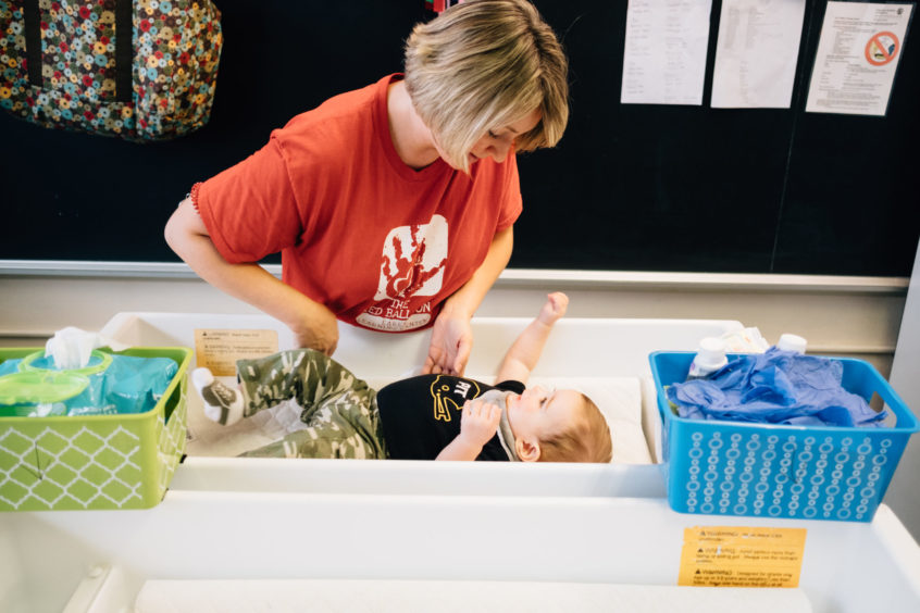 Image: An early learning professional changes the diaper of a young child who is placed on a changing table. The young child looks up at the caregiver while raising their hands in the air.