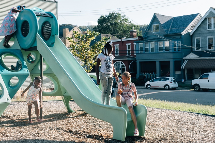 Image: Three young children play together on a green slide at their local playground.