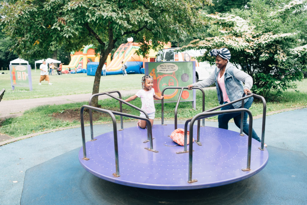 Image: A young child and her caregiver play together joyfully at a local playground, spinning together on a Merry-Go-Round.