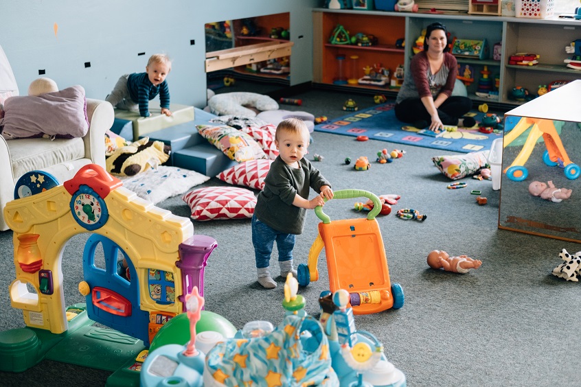Image: An early childhood professional sits on the floor, supervising two toddlers who are playing with several types of colorful toys.