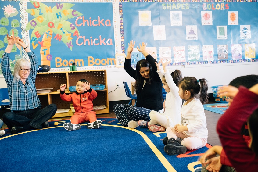 Image: Early learning professionals sit in a circle with young children, all raising their hands and smiling happily.
