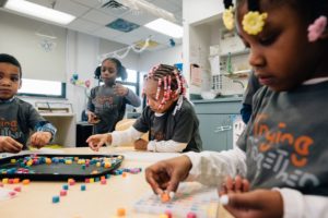 Image: Four young children play together in an early childhood classroom. Three of them sit at a table playing with small colorful blocks. One child stands beside the table watching the others play.