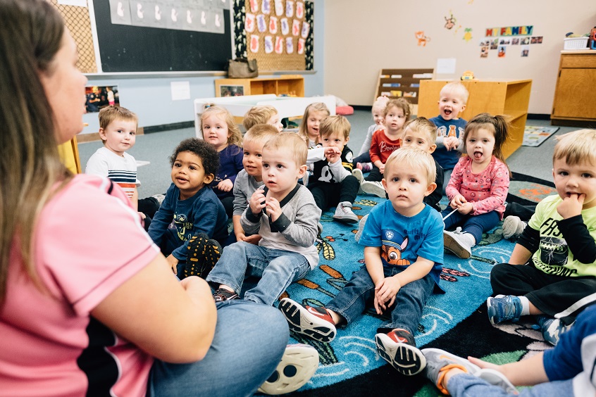Image: A group of young children sit in a group together on the floor in front of an early learning professional. Several children are grinning widely.