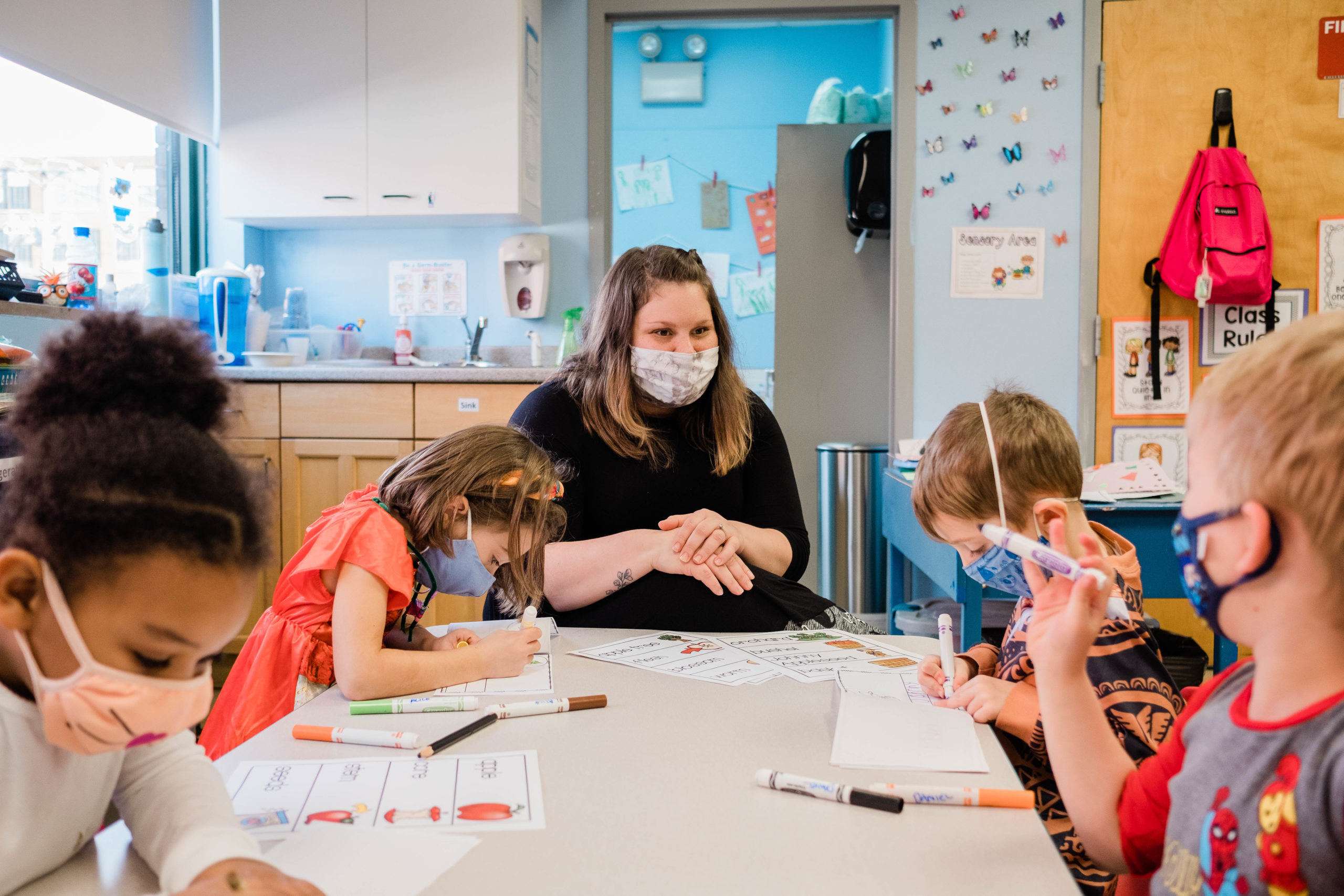 teacher wearing face mask teaches a group of students wearing face masks as a table