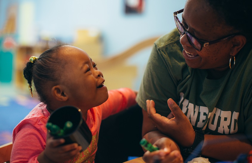 Young Black girl and caregiver smile as they look at each other and color with green markers.