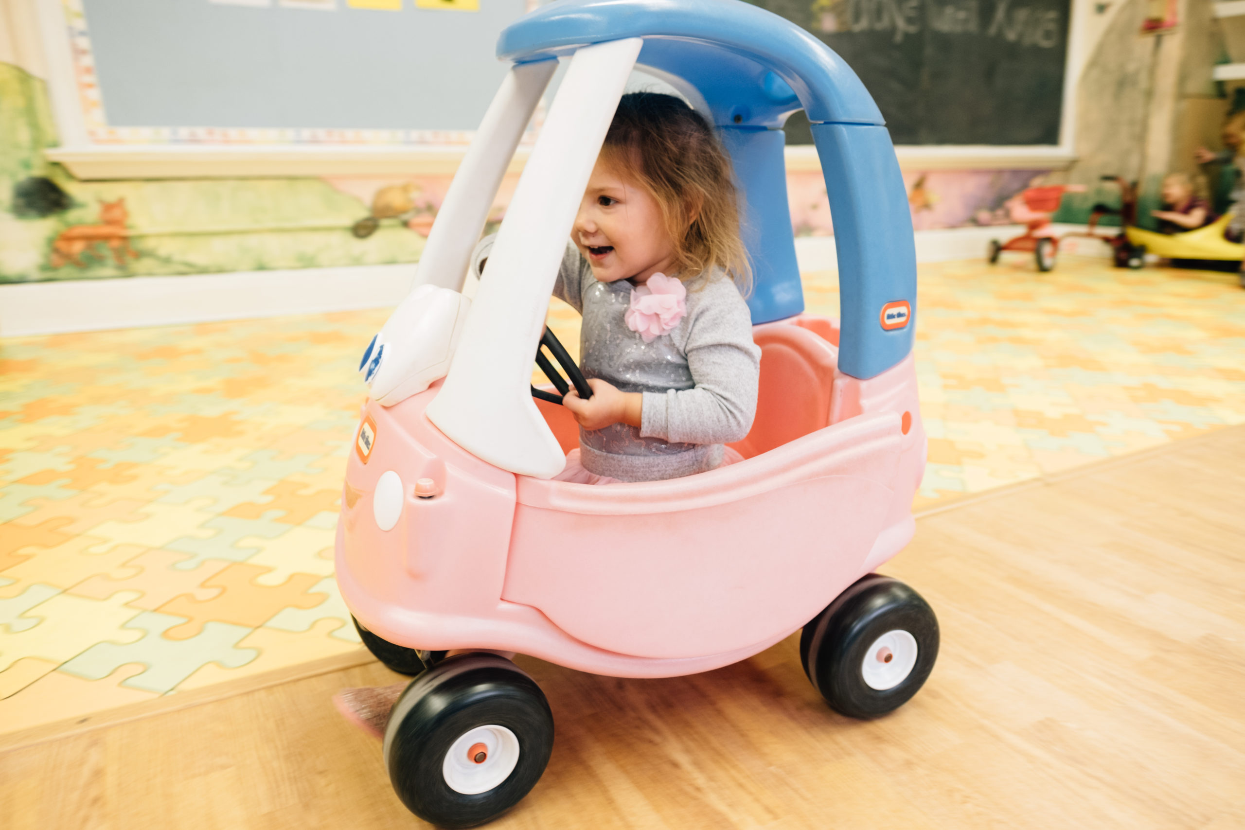 A young toddler riding in a pink and purple toy car.
