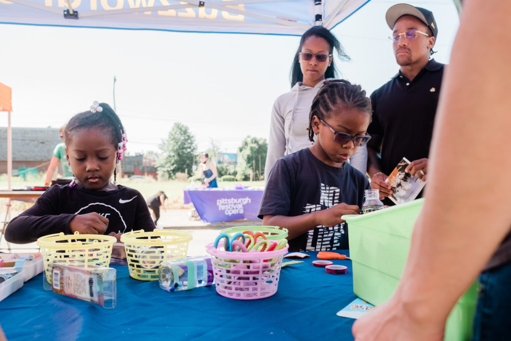 Two young children make a craft at a table while their parents watch from the back