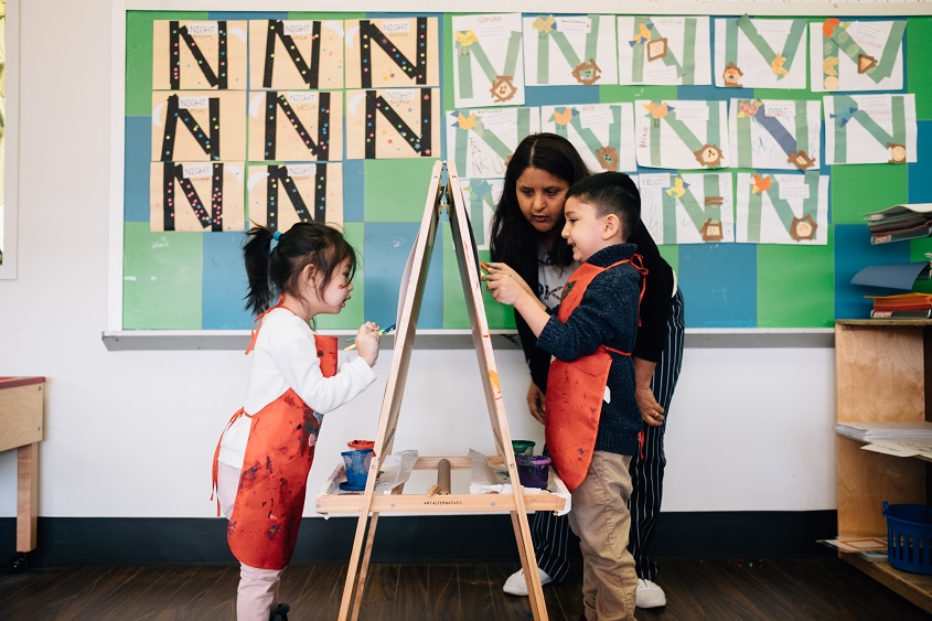 Caregiver in classroom with two children on opposite sides of an art easel.