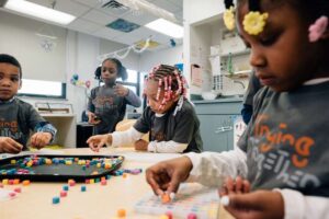 Anti-Racism Tools | Image: Four young children play together in an early childhood classroom. Three of them sit at a table playing with small colorful blocks. One child stands beside the table watching the others play.