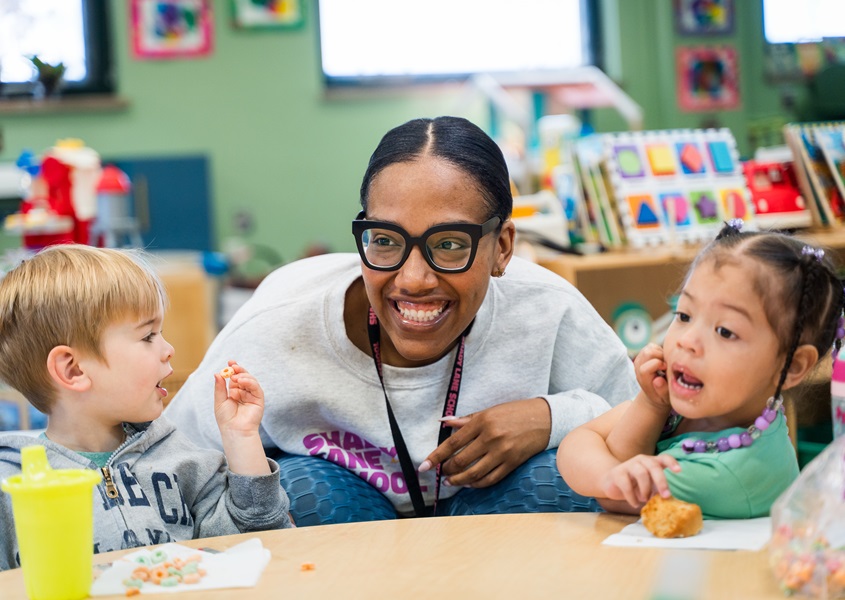 Black female early educator crouches down between two toddlers at table, one a white boy with blonde hair to her right, the other a Black girl with braids to her left.