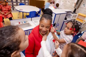 Adult woman interacts with two young children in preschool classroom